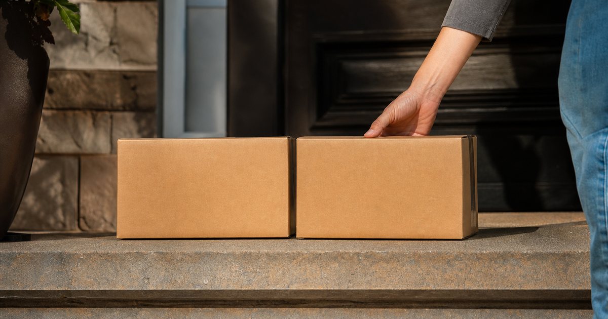 Two identical brown cardboard parcels sitting side by side on the front step of a suburban house, afternoon sun, a person's hand reaching for one of them, no visible labels or text